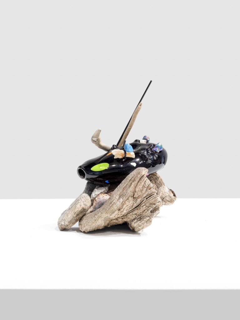 Glossy black sculptural form with neon green and blue marks, pierced by sticks and adorned with crystals and shells, balanced on a rough, woodlike piece on a white platform.