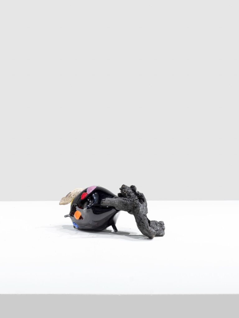 Small glossy black sculpture with rounded form, dotted with small red, pink, orange, and blue shapes. An irregular, textured gray branch pierces through, resting on a stark white platform.