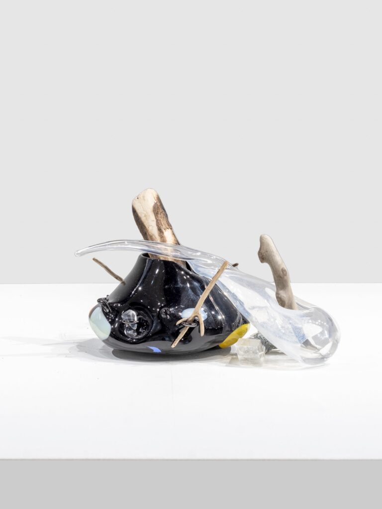 Sculpture of glossy black and transparent glass forms with smooth, rounded shapes, pierced by rough wooden branches and twine, resting on a white surface with scattered textured blocks.