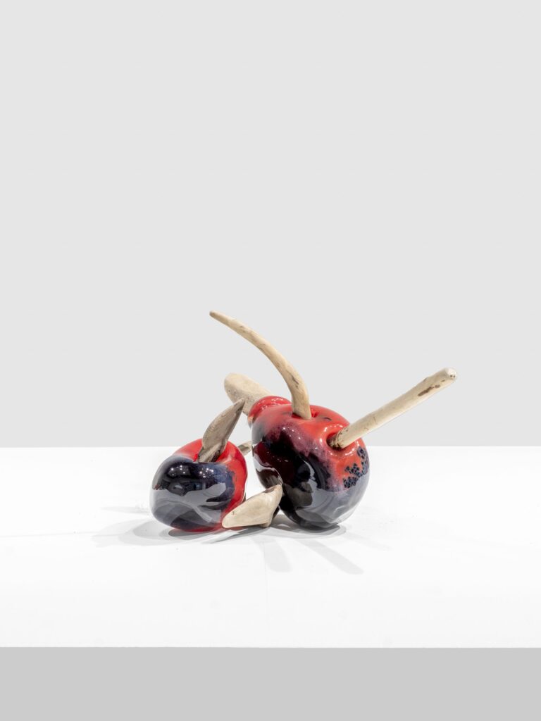 Glossy red and black ceramic forms with rounded shapes and smooth surfaces, pierced by curved, rough-textured wooden sticks, arranged in a dynamic, leaning composition on a white platform.