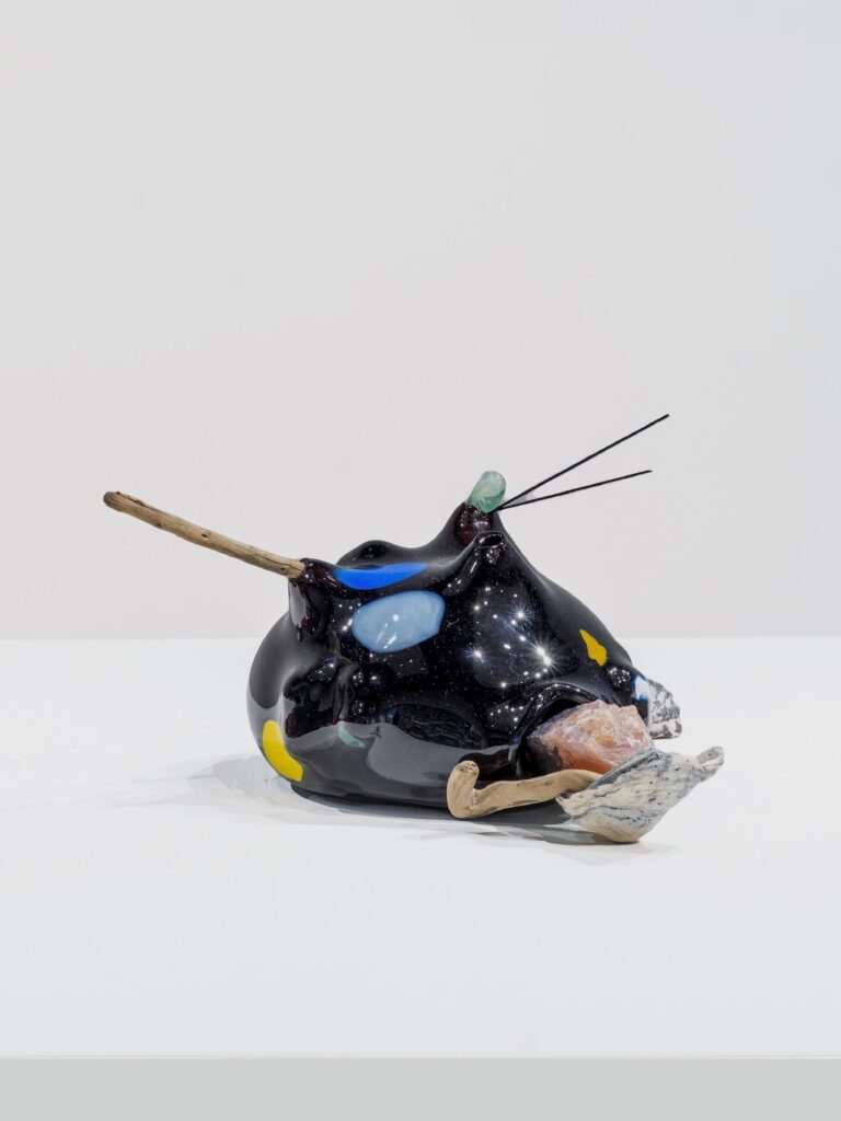 Glossy black sculptural form with a starry surface, marked with blue, yellow, and aqua shapes. Sticks, stones, and crystals emerge from openings, arranged on a white display platform.