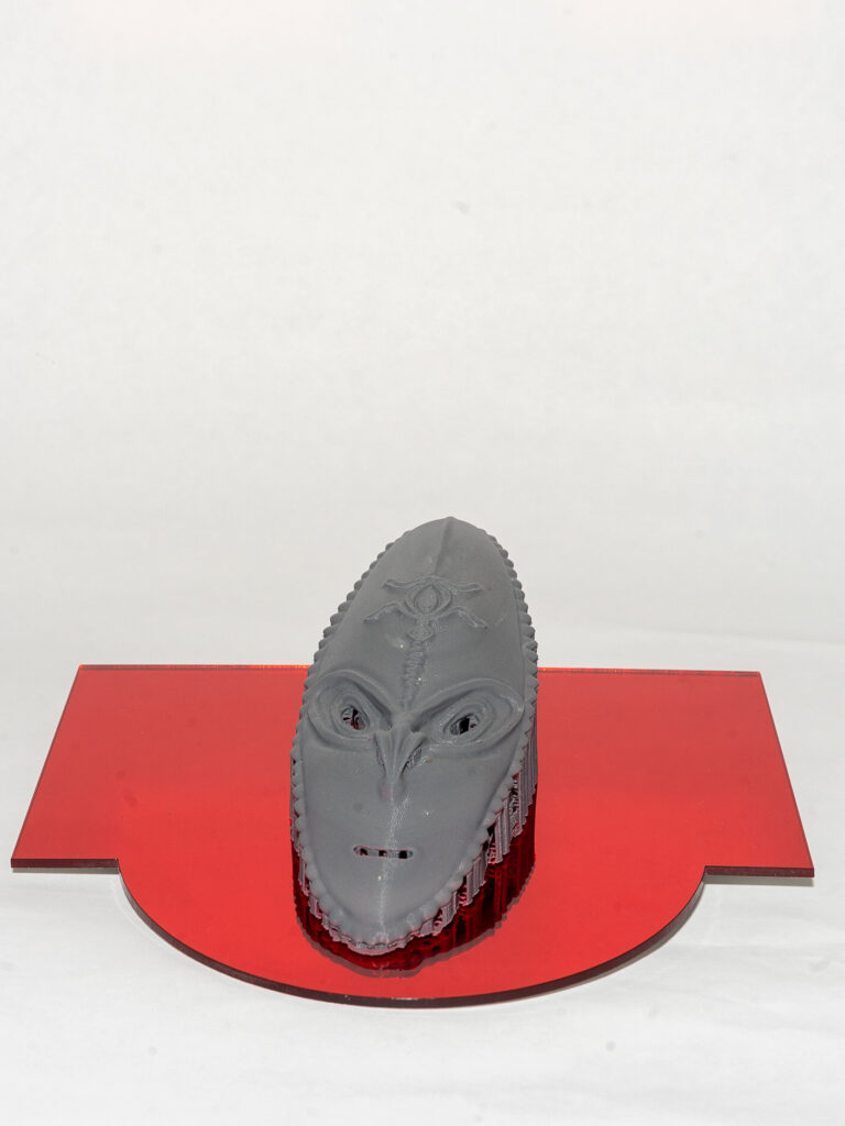 A grey, oval mask sits on top of a red geometric shape. The mask includes stylized eyes, nose and mouth, with an animal shape on the forehead.