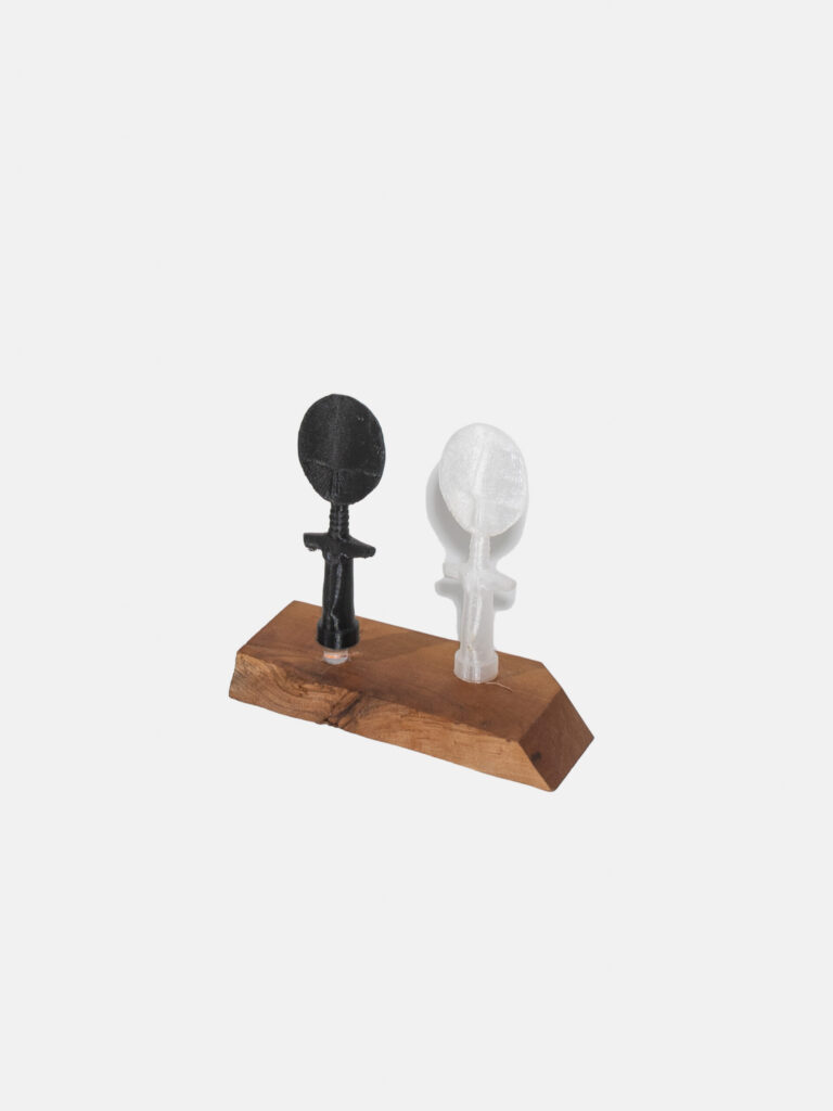 Two 3D humanoid figures, one black, one white, stand on a wooden block. The figures have simplified bodies and large heads, with simple outlines of facial features.