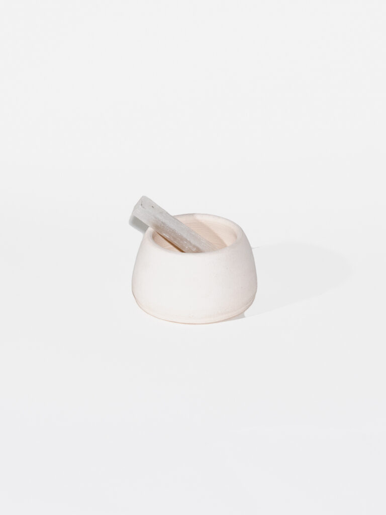 An off-white bowl sits on a white background with a rectangular crystal propped inside. The crystal sticks out of the bowl like a spoon.