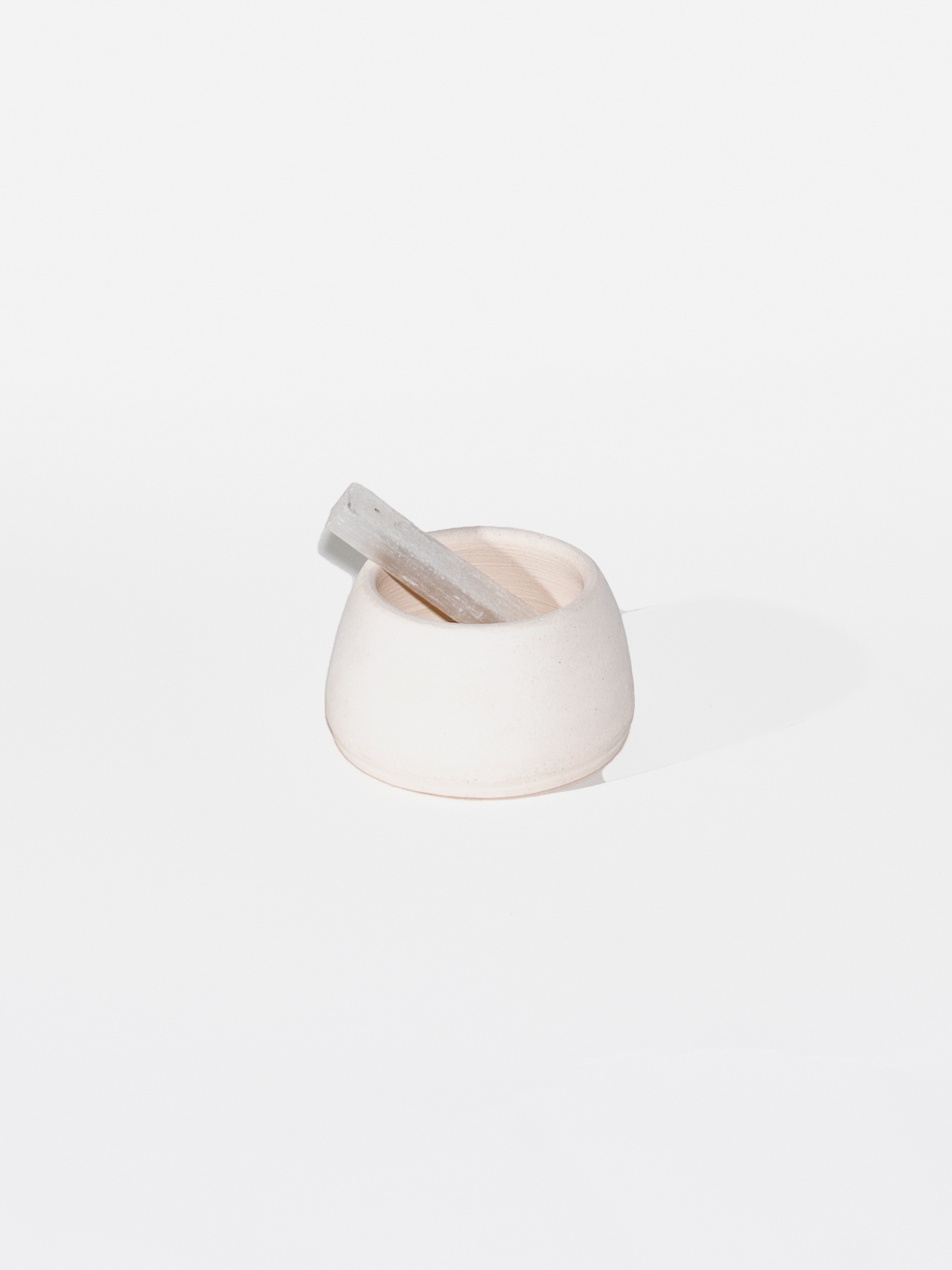 An off-white bowl sits on a white background with a rectangular crystal propped inside. The crystal sticks out of the bowl like a spoon.