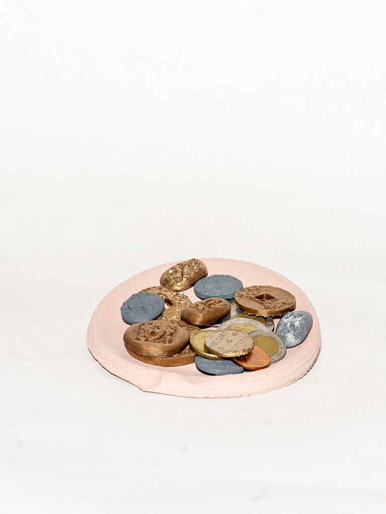 Gold and silver colored coins sit on a pale pink circular tray in front of a white background. The coins vary in shape and size with different engravings.