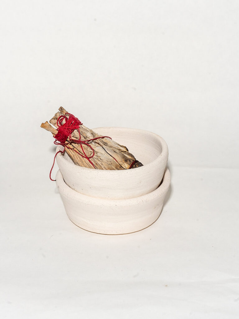 Two ceramic bowls are stacked on top of each other on a white background. A bundle of dried sage tied together with red string sits in the top bowl.