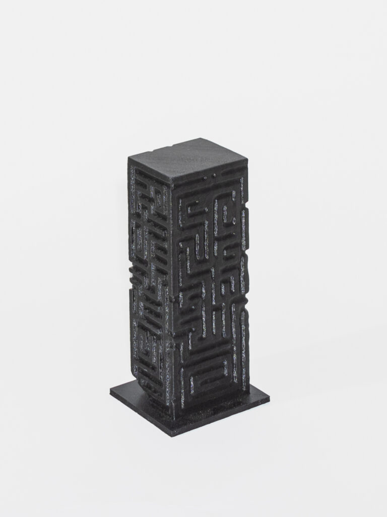 A dark grey rectangular column with two sides visible sits on a grey square. The column is engraved with geometric patterns and is in front of a white background.