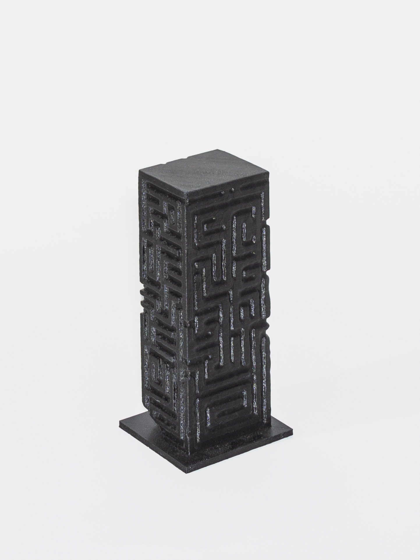 A dark grey rectangular column with two sides visible sits on a grey square. The column is engraved with geometric patterns and is in front of a white background.