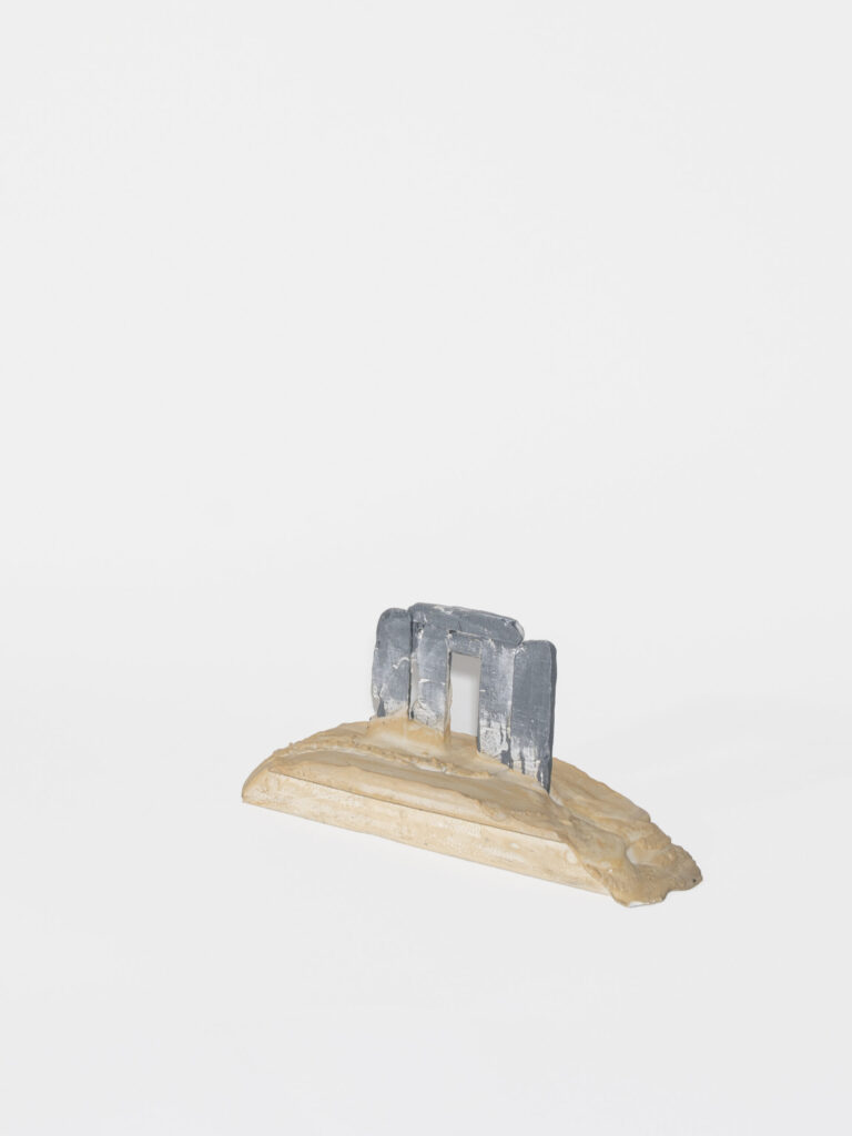 A grey form emerges from a semi-circular beige organic shape. The form is made up of four vertical columns topped by two horizontal columns.