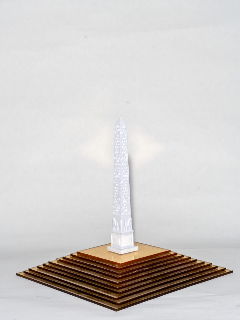 A white obelisk is engraved with small shapes. It sits on top of a brown square with stairs visible on two sides leading up to the obelisk.
