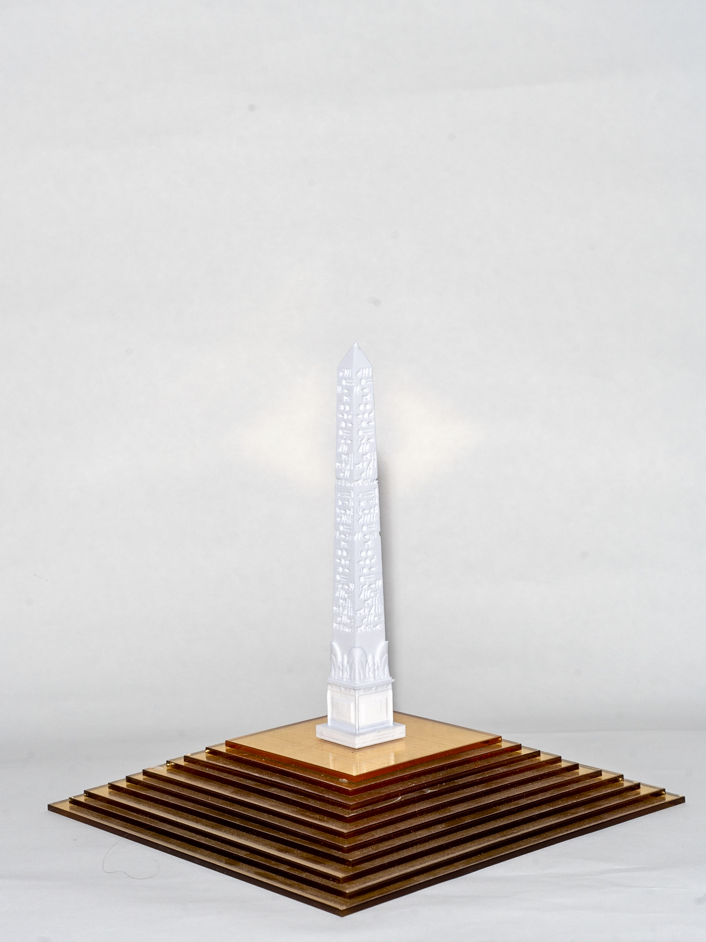 A white obelisk is engraved with small shapes. It sits on top of a brown square with stairs visible on two sides leading up to the obelisk.