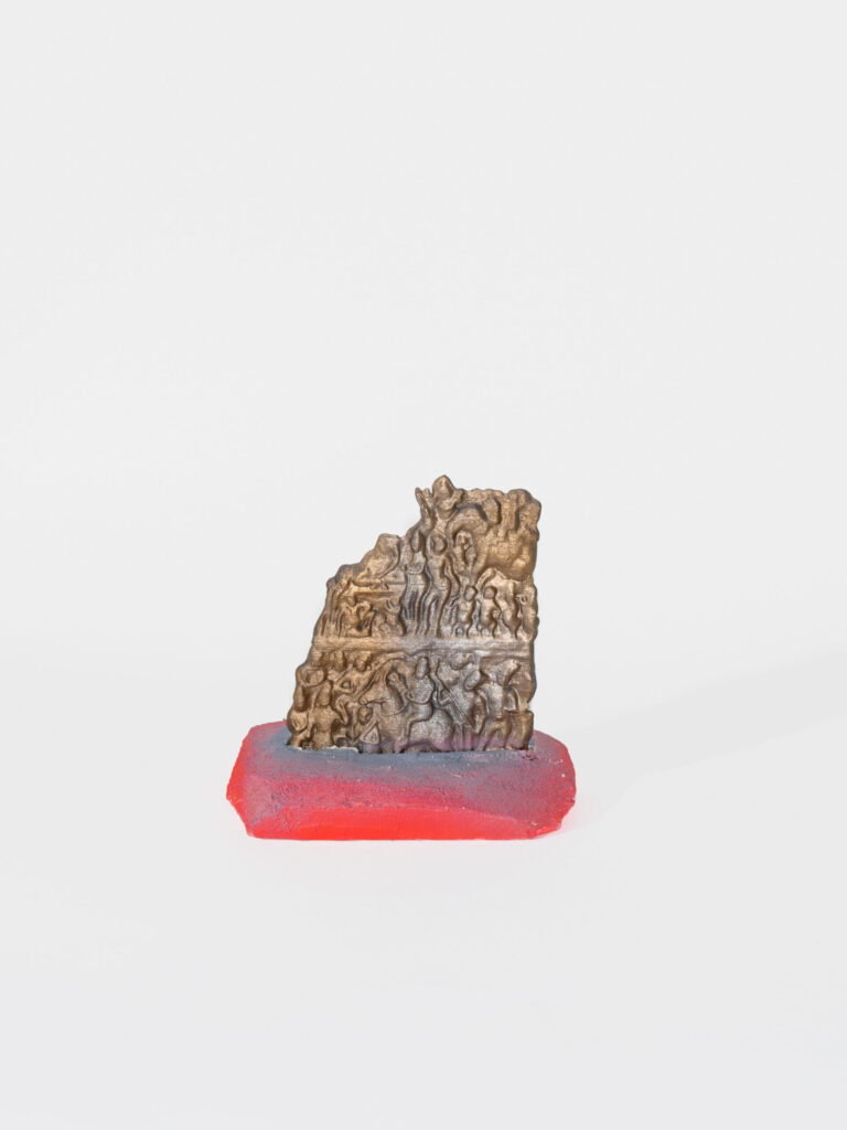 A 3D gold organic form with details of people, some on horseback, others dancing. The form sits on a red and grey stand that is vaguely rectangular.