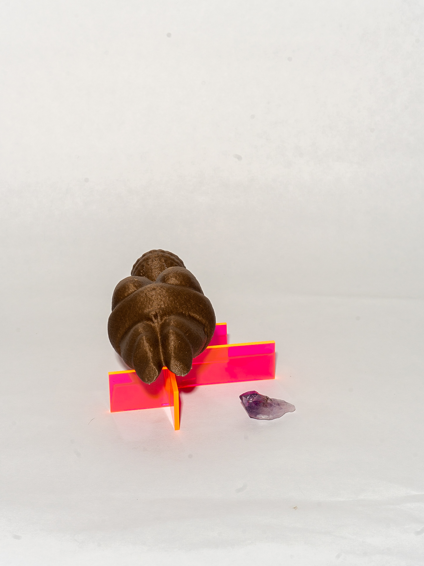 Three objects: a purple melting gemstone, a neon pink 3D form made of horizontal and vertical lines, and a brown humanoid figure lying on the pink form.