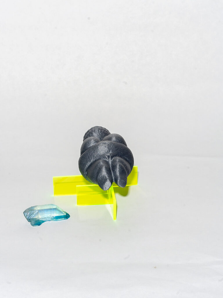 Three objects: a blue melting gemstone, a neon yellow 3D form made of horizontal and vertical lines, and a grey humanoid figure lying on the yellow form.