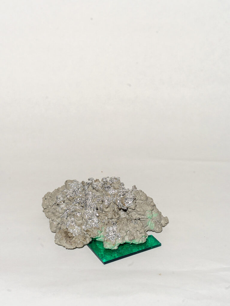 An abstract 3D form rests on a reflective green square. The form is grey with reflective silver parts on the top.