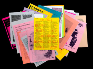 A stack of colorful printed papers, including yellow, pink, orange, and gray sheets, with text, images, and drawings spread out on a black background.