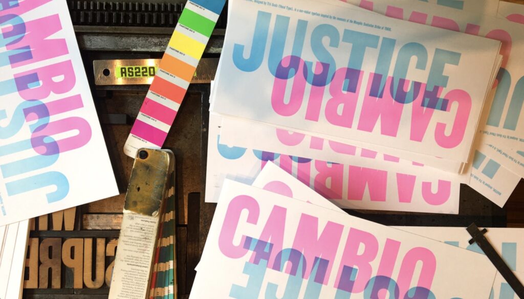 Stacks of printed posters with large text overlapping, “JUSTICE” in blue and “CAMBIO” in pink, placed beside a colorful paint swatch card and metal printing tools.