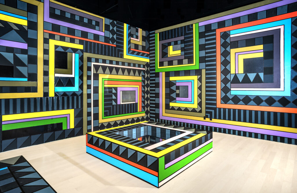 Installation view of two walls covered entirely with geometric black and blue stripes and bright outlines of rectangles. A similar pattern covers a rectangular structure on the floor.