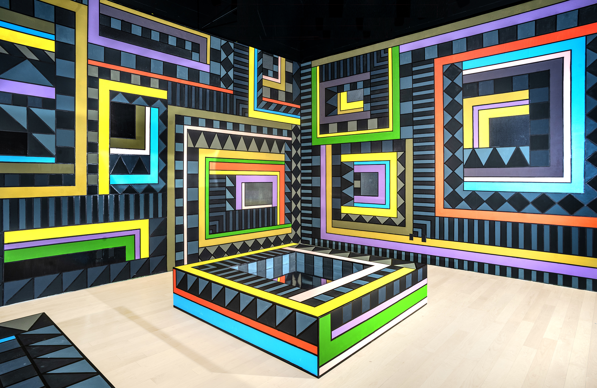 Installation view of two walls covered entirely with geometric black and blue stripes and bright outlines of rectangles. A similar pattern covers a rectangular structure on the floor.