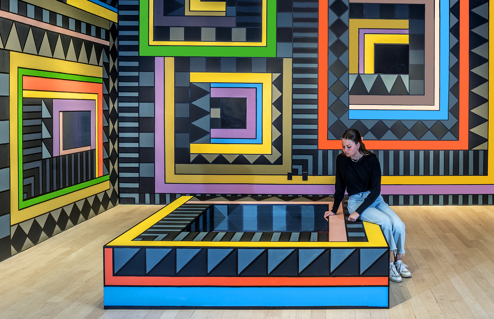 Installation view of a wall covered in geometric patterns consisting of primarily black, blue, and grey, with bright rectangular pops of color. Similar patterning covers a rectangular structure on the floor, which a young woman sits on.