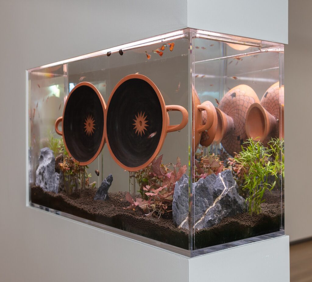 An aquarium built into a gallery wall containing small orange fish, live plants, and dark substrate. Inside the tank, two terracotta vessels with black interiors and sun motifs are submerged.