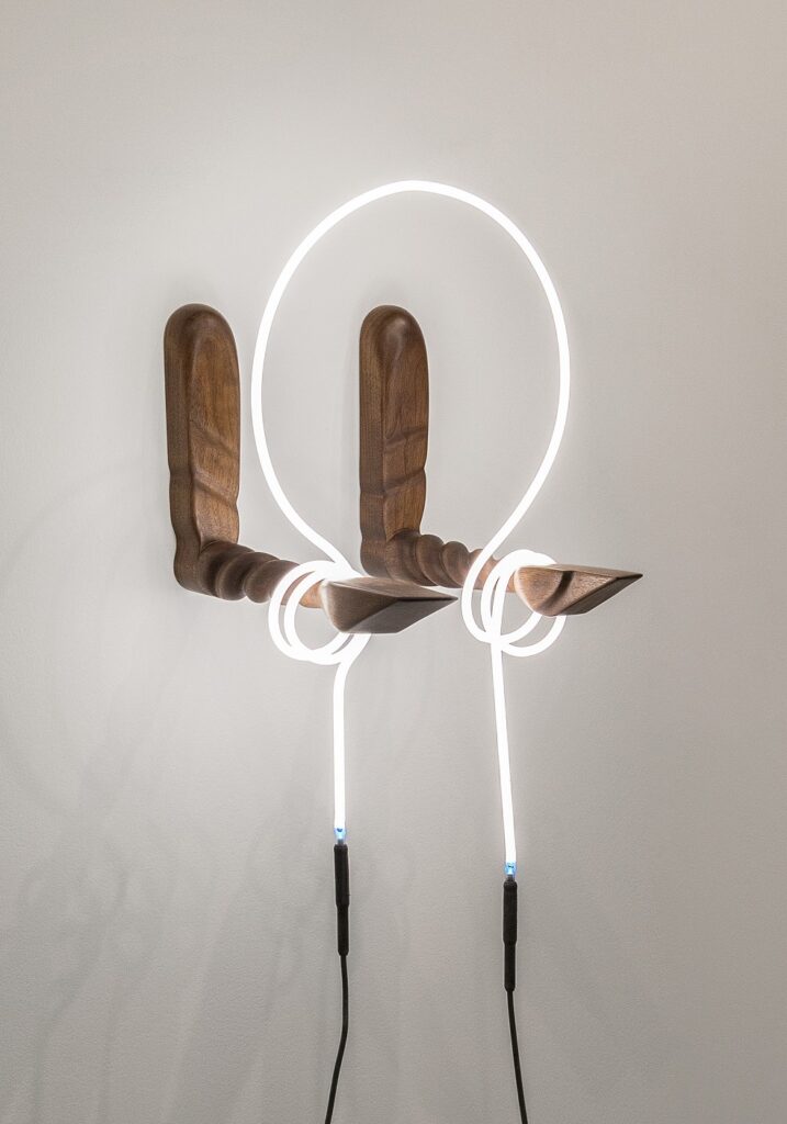 Two wooden arms are mounted to a white wall and extend outward like arms at a 90-degree angle. Fluorescent tubing wraps around both arms and rises into a circular shape.