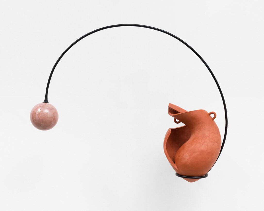 An abstract sculpture consisting of an orange ceramic vessel. A black steel line curves upwards, connecting the bottom of the vessel to a pink marble sphere to the left.