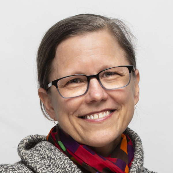 Portrait of Carol Stakenas, a light-skinned woman with glasses who is wearing a multicolor scarf and gray collared sweater or jacket.