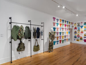 Installation view showing muted clothing hanging on clothing racks, and a mannequin dressed in camouflage. On the right, repeated images of a monkey's head in different colors cover the wall.