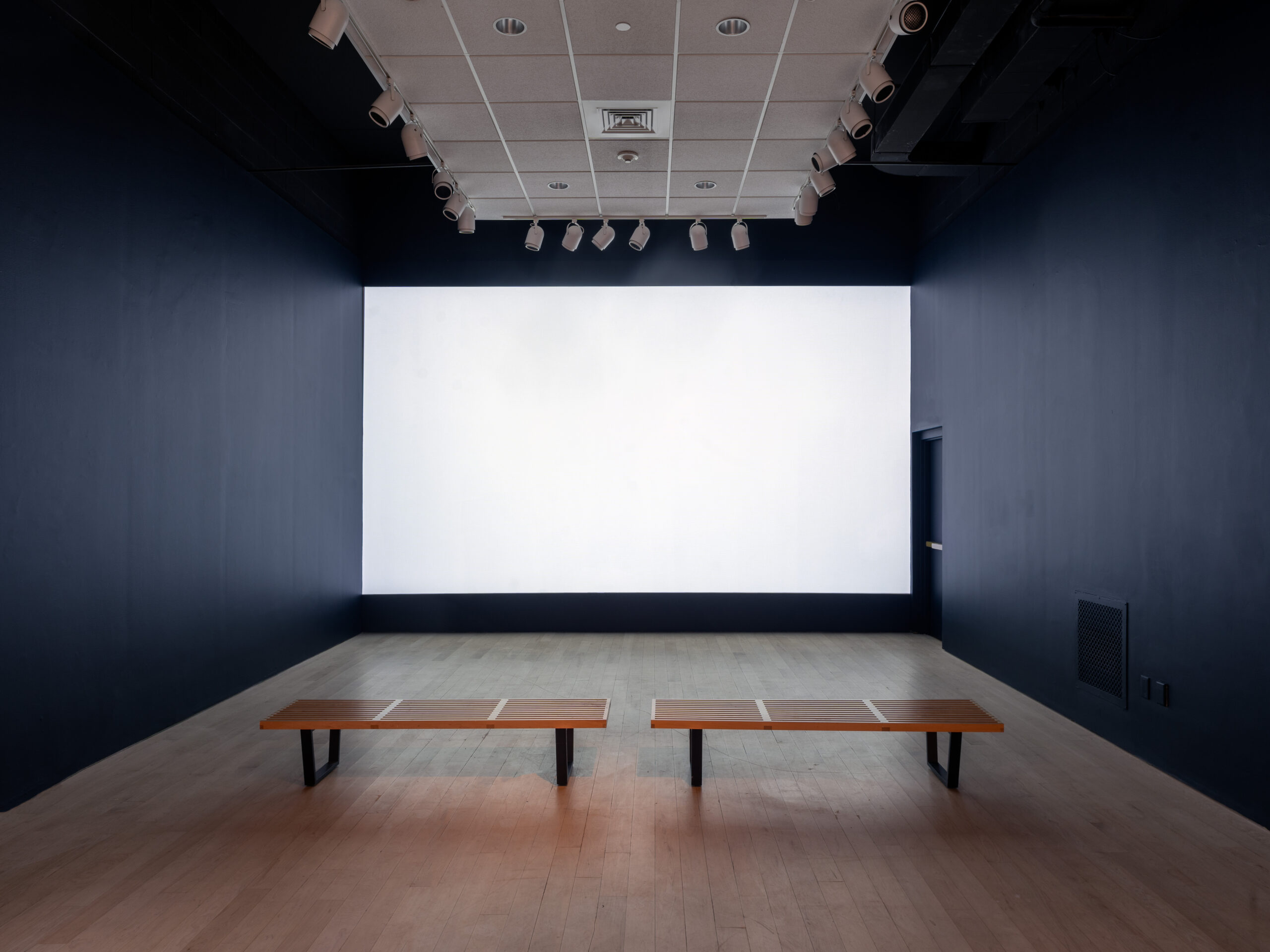An installation view of a gallery with two benches facing a blank white screen. The rest of the walls on either side are completely black.