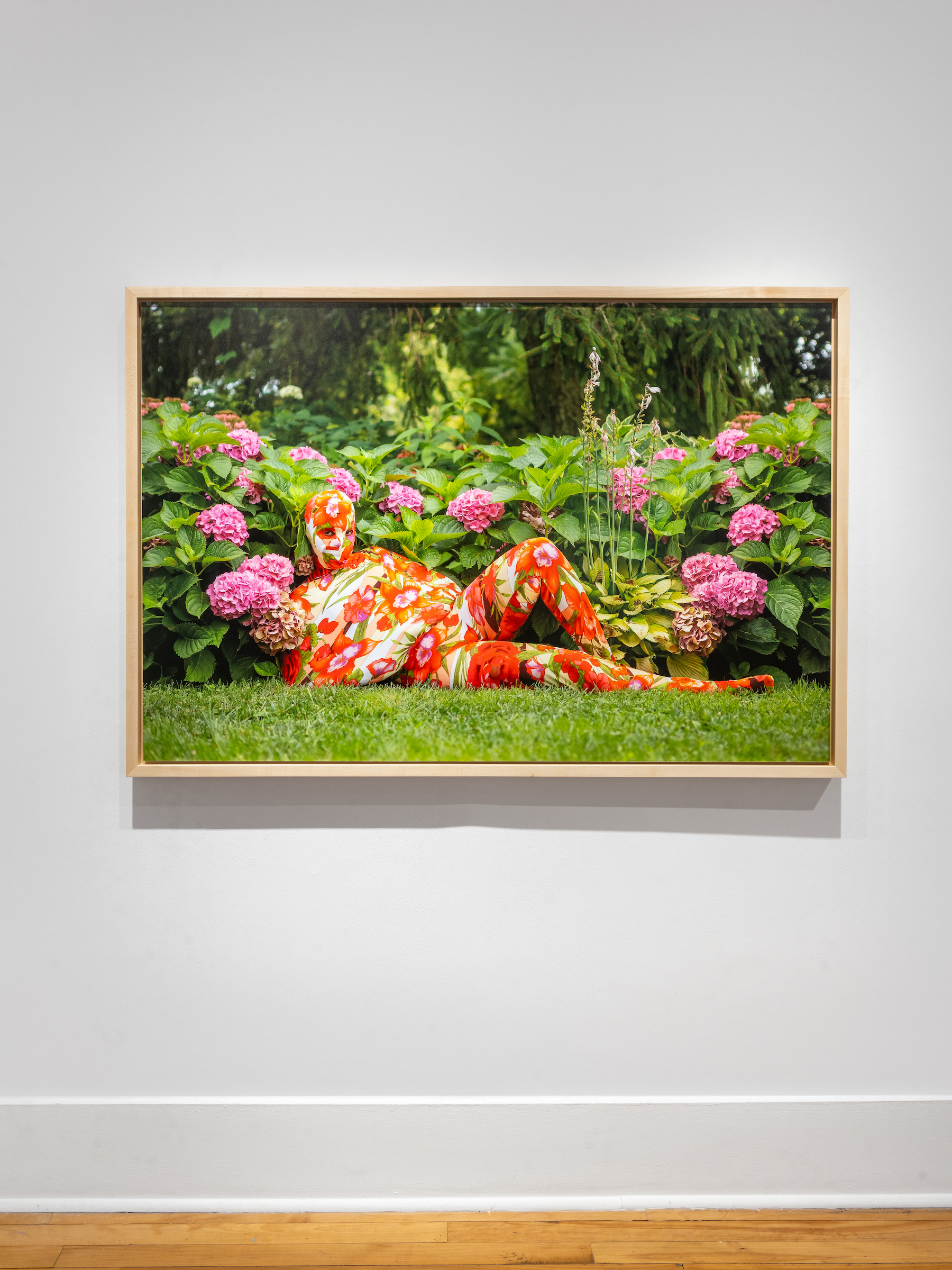 Lying in the grass among blooming pink flowers, a figure is clad head to toe in a red, purple, and green floral ensemble that covers their body.