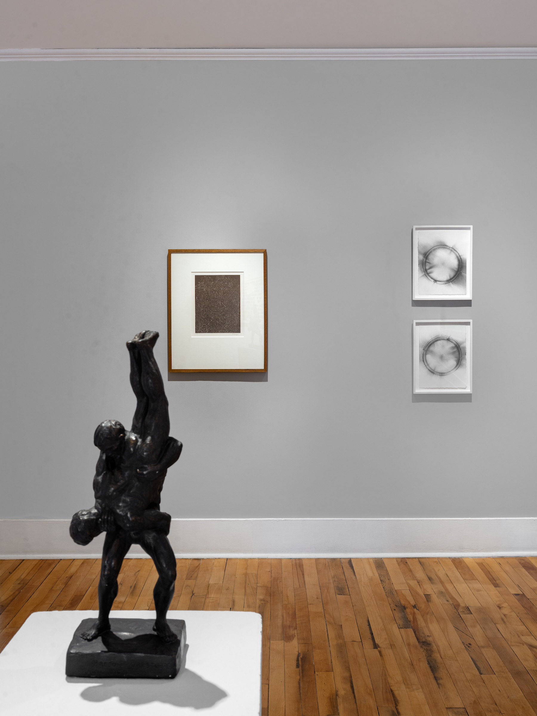 An installation view with a sculpture of two figures wrestling in the foreground. Behind is a larger artwork nex to two stacked smaller artworks hung on a grey wall.