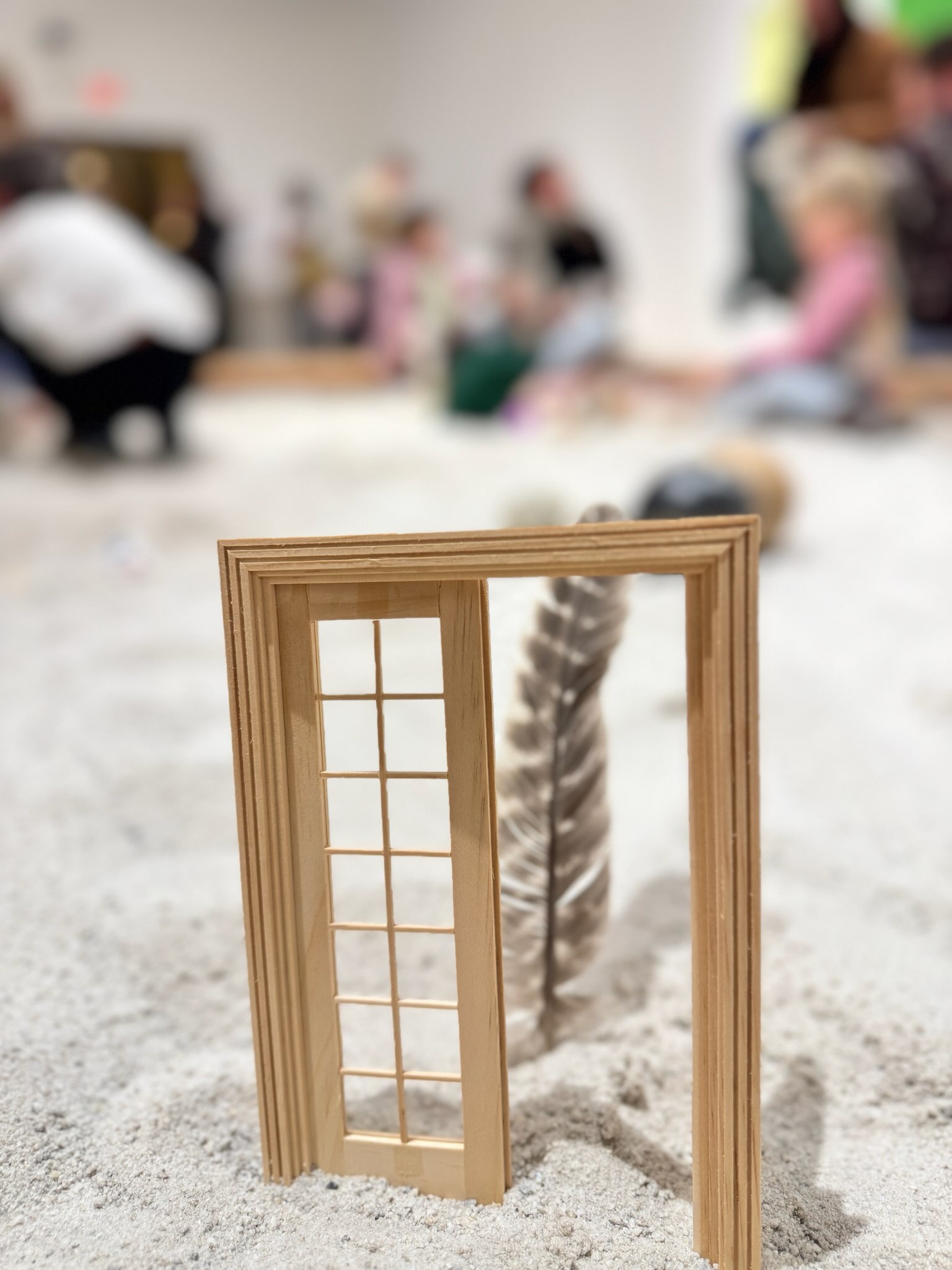 A miniature door frame protruding from the sand with an opening in it showing a brown and white striped feather also sticking out of the sand.