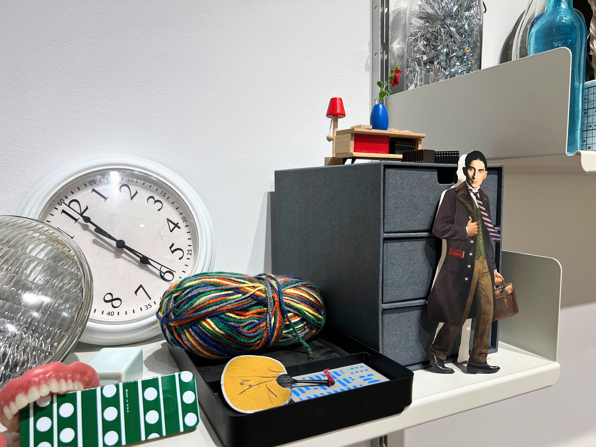 A white shelf against the wall holding an assortment of objects, including a multi-color ball of string, a storage shelf, dollhouse furniture, a clock, a cutout of Franz Kafka, and a fake set of teeth.