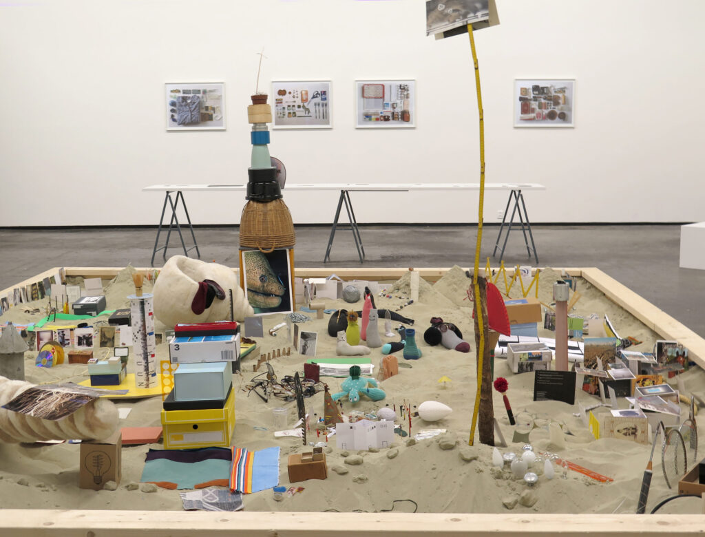 Art exhibition featuring various colorful small objects filling a sandbox. Some are stacked on top of each other, and some are buried in the sand.