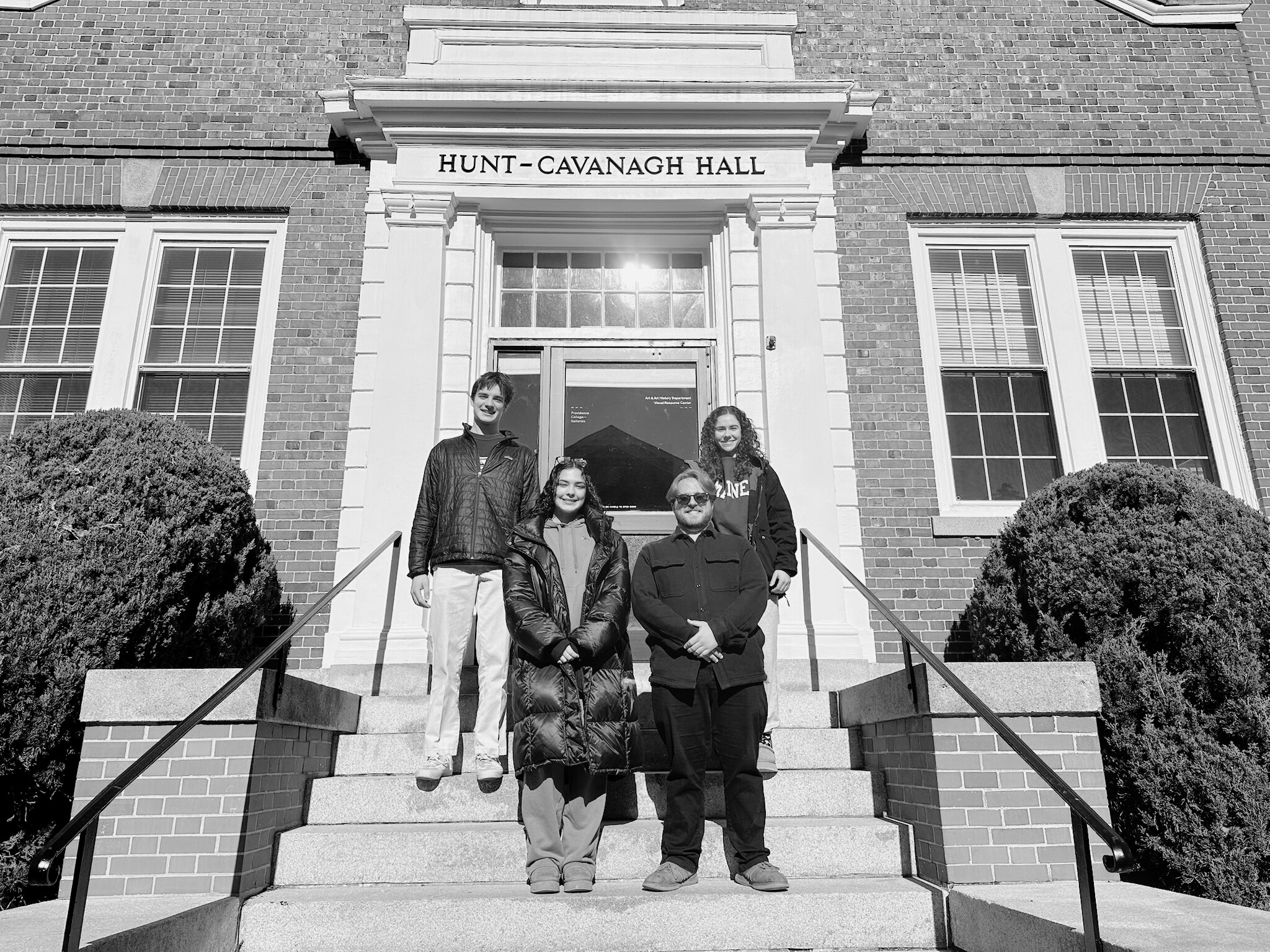 Four students in winter coats stand on the front steps of Hunt-Cavanagh Hall at Providence College.