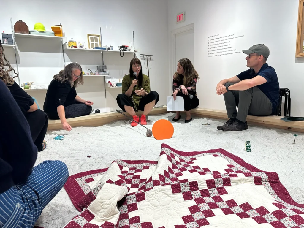People sitting around a very large sandbox, indoors.